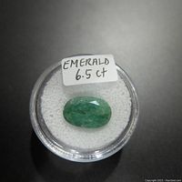 Oval emerald gemstone in a clear round display container with a white foam base, label reads 'EMERALD 6.5 ct'.
