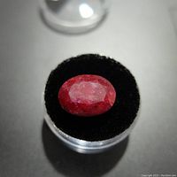 Top view of the loose oval-shaped red ruby gemstone showing color and surface texture on a black velvet cushion inside a clear container.