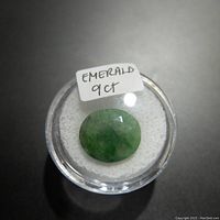 Oval cut green emerald gemstone approximately 9 carats in weight placed in a clear round container labeled 'EMERALD 9CT'.