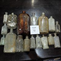 Top view showing the entire collection of 17 various old glass bottles arranged on a dark surface with maxsold lot number card