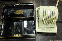 Metal cash box with compartments alongside cream-colored Paymaster Ribbon Writer counting machine on a dark surface.