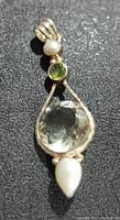Close-up photo of the silver pendant showing the large pale green faceted stone, smaller green peridot, and two pearls on a textured dark surface.