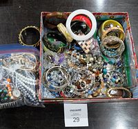 Photo showing a box with assorted vintage fashion jewelry including multiple bangles, bracelets, and necklaces in various colors and materials.