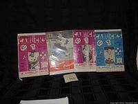 Photo showing 4 of the Vancouver Canucks 1963-64 WHL hockey programs arranged on black fabric. Programs have pink and blue covers with vintage player images and logos.