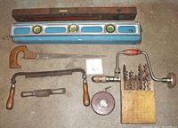 Photo showing wooden Stanley level, metal Stanley level in wood case, keyhole saw, hand brace with drill bits, tape measure and draw and spoke planes.