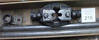 Top view of metal pipe threading die head showing clamps with size markings and threading dies attached to handle bar, placed on a wooden surface inside case.