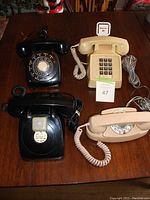 Top view of four vintage telephones on a wooden table showing two black rotary phones, one beige rotary phone, and one beige push-button phone.