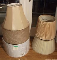 Photo showing five large lamp shades stacked and arranged on floor near window.