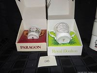 Two loving cups in their original boxes, one Paragon on the left with gold handles and floral motif, one Royal Doulton on the right, white with commemorative text and green floral design.