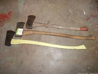 Three axes laid on the ground showing full length and handles with the yellow handle axe on bottom, natural wood handle axe in middle, and shorter natural wood handle axe on top.