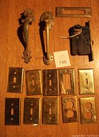 Photo showing the two vintage door handles, brass mail slot, black metal door lock, and multiple brass switch plates arranged on wood surface.