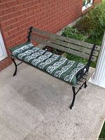 Outdoor bench with cast iron ends, wood slats, and green/white patterned cushion on cement patio against red brick wall and white column
