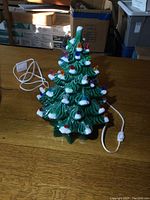Front angled view showing plugged in ceramic Christmas tree with colorful bulb lights and attached white power cord with switch on a wooden surface.