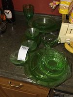 Green Depression glass collection displayed on a kitchen counter, including large serving plates, bowls, and a green goblet, showing vintage condition and transparency.