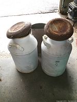 Two white steel milk cans side by side, each with rusted lids and metal handles, showing signs of wear and rust.