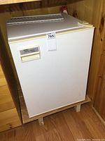 Front angle view of the white Danby Diplomat mini fridge sitting on wooden floor inside a wooden space.
