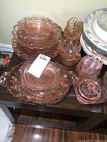 Multiple pink depression glass bowls, plates, and serving plates stacked on a table showing ribbed and starburst patterns with some chips.