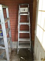 Front view of the aluminum 6 ft step ladder leaning against a brick wall, showing all steps and top platform that has paint splatters.