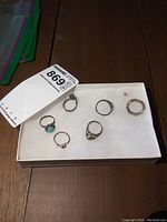 Six sterling silver rings displayed in a white box on a wooden surface, showing variety of styles, stones, and sizes.