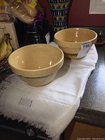 Pair of beige stoneware mixing bowls displayed on white cloth on countertop alongside kitchen appliances and bananas. The bowls have a smooth glossy interior and textured exterior with decorative patterns.