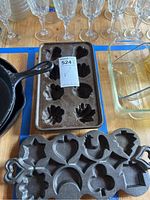 Shows a dark cast iron cookie pan with nine cavities in varied shapes including stars, hearts, and pumpkin-like figures, and an 8-cavity cast iron muffin pan in the background along with glassware and a clear glass baking dish.