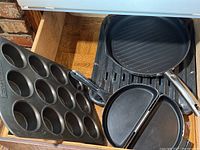 Photo showing black metal muffin pan with 12 slots, round grill pan with ridges, divided omelette pan with two compartments, and broiler tray under them inside a wooden drawer.
