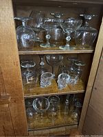 View of various clear crystal and glass stemware and tumblers inside a wooden cabinet, arranged on three shelves.