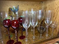 Various wine glasses including red wine glasses, champagne flutes, goblets, and a small clear shot glass displayed on shelf