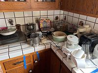 Overview of kitchen countertop showing various kitchen appliances and baking items including Cuisinart food processor, Krups appliance, cake keeper, toaster, wire racks, ceramic bowls, and more