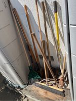 Photo showing a collection of long handle outdoor tools leaning against a wall outside including rakes, shovels, and a post hole digger