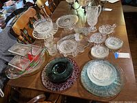 Photo showing a collection of various clear glass and crystal serving ware including platters, plates, cups, pedestal cake plates, bowls, and ashtrays arranged on a wooden table.