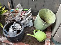 Photo showing multiple open bags of soil inside and around a light brown cracked terra cotta pot, next to a large pale green metal bucket and a small green watering can on brick ground outdoors.