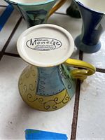 Close-up of bottom of ceramic cup showing brand mark "Elizabeth Murray Montage" along with dishwasher safe and stoneware designation, cup inverted to show detail.