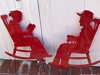 Pair of red metal silhouettes of a man and woman seated in rocking chairs, shown together against a wood panel backdrop, placed on a brick surface, displaying weathering and wear