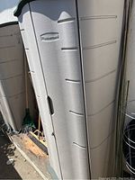 Front view of the Rubbermaid outdoor storage cabinet showing textured beige plastic construction and dark green top with the Rubbermaid logo.