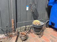 Bicycle-style black metal plant holder on wheels containing a large pot with cactus, next to small iron pot and rectangular plant shelf on ground