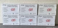 Six new TCP energy efficient compact exit signs in packaging, showing front label with product details, 'EXIT' text, and suitability for damp locations.