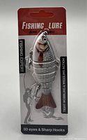 Front view of packaged fishing lure showing the segmented body, 3D eyes, hooks, and branding