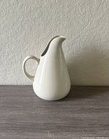 Image of vintage Russel Wright signed stoneware pitcher in linen color on wooden surface against neutral wall.