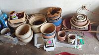Large assortment of garden pots of various sizes and colors, wooden flower box, and small rusty wheelbarrow with dust in a garage setting.