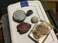 Photo of geodes and mineral specimens on white tote lid, showing varied shapes and colors including polished sliced geode, green crystal, reddish mineral chunk, and a box with crystal clusters.