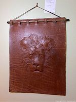 Full view of the rectangular leather wall hanging showing the embossed lion's head and hanging structure with wooden dowel and leather loops.