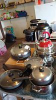 Wide view of the kitchen counter showing pressure cooker, several pots and pans including cast iron, aluminum, nonstick pans with glass lids, and a red and silver stacked kettle.
