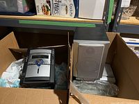 Photo showing main sound system unit and one speaker packed in a cardboard box surrounded by packing material.