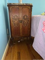 Full front view of the wooden stand showing marble top, door with floral inlaid wood panels, curved legs and ornate metal handles.