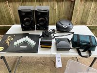 Lot of Polaroid camera, two Sony speakers, Sony CD player, photo printer, manuals, and camera bag on a white table outdoors