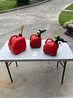 Photo of three red gas cans on a folding table outdoors: two 1-gallon cans and one 2-gallon can with different colored spouts.
