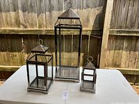 Set of three glass lanterns with metal frames of different sizes on a white table with wooden fence background.