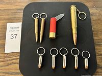 Top-down photo of 8 keychains including 5 bullet-shaped keychains, 2 bullet-shaped keychains with ring, 1 shotgun shell-shaped keychain with a small knife.