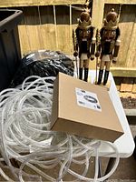 Two wooden nutcrackers, clear LED rope light coiled, black string lights in packaging, and boxed tree skirt on outdoor table.